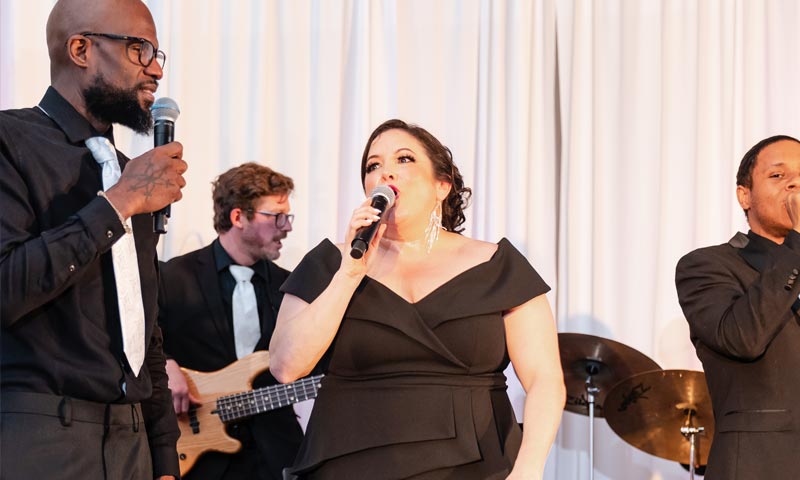 Amy Yassinger Orchestra Performing with 4 singers, background has man playing keyboard, LED wall, person playing drums.